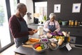 Senior biracial couple in aprons, cutting and peeling vegetables and using tablet in kitchen at home