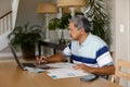 Senior biracial man doing paperwork and using laptop in dining room