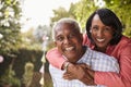 Senior black couple piggyback in garden looking at camera