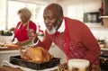 Senior black man basting roast turkey in preparation for Christmas dinner, his wife chopping vegetables in the background, close u