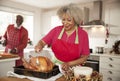 Senior black woman basting a roast turkey in preparation for Christmas dinner, her husband chopping vegetables in the background,