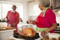 Senior black woman preparing a roast turkey for Christmas dinner turns to talk to her husband, chopping vegetables in the backgrou Royalty Free Stock Photo