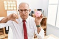 Senior business man holding piggy bank with angry face, negative sign showing dislike with thumbs down, rejection concept Royalty Free Stock Photo