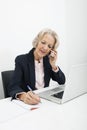 Senior businesswoman writing in book while answering cell phone at desk in office Royalty Free Stock Photo