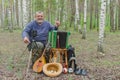 Senior camper is having rest in birch forest, sitting on a wicker stool and holding mandolin
