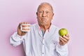 Senior caucasian man holding denture and green apple puffing cheeks with funny face