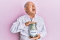 Senior caucasian man holding jar with savings angry and mad screaming frustrated and furious, shouting with anger looking up Royalty Free Stock Photo