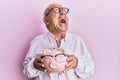 Senior caucasian man holding piggy bank with glasses angry and mad screaming frustrated and furious, shouting with anger looking Royalty Free Stock Photo