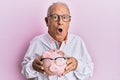 Senior caucasian man holding piggy bank with glasses in shock face, looking skeptical and sarcastic, surprised with open mouth Royalty Free Stock Photo