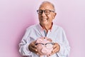 Senior caucasian man holding piggy bank with glasses winking looking at the camera with sexy expression, cheerful and happy face Royalty Free Stock Photo