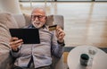 Senior cheerful handsome man lying down on sofa or couch at his home or office using hand held tablet device and credit card for Royalty Free Stock Photo