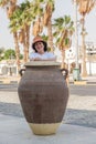 Senior citizen brunette woman in wicker hat stands behind a large clay pot on the street