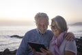 A senior couple embrace near the beach while looking at the tablet together. Sitting on the cliffs, twilight, two elderly people Royalty Free Stock Photo