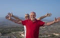 Senior couple of friends people standing on hill with mountain and city on background looking at camera with arms raised - active Royalty Free Stock Photo