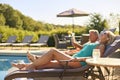 Senior Couple On Holiday With Woman Wearing Wireless Earbuds And Man Reading Book By Swimming Pool Royalty Free Stock Photo