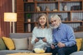 Senior couple playing video game console with joysticks sitting on couch in living room. Funny Mature people family having fun Royalty Free Stock Photo