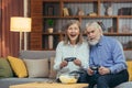 Senior couple playing video game console with joysticks sitting on couch in living room. Funny Mature people family having fun Royalty Free Stock Photo