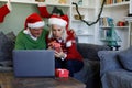 Senior couple in santa hat sitting on couch opening a gift box while having a video call on laptop a Royalty Free Stock Photo