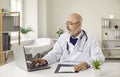 Senior doctor in glasses and white medical gown working on laptop sitting at workplace in office. Royalty Free Stock Photo