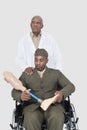 Senior doctor with US military officer holding artificial limb as he sits in wheelchair over gray background