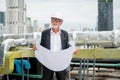 Senior engineer man hold drawing of building and look to his right side during work on rooftop of construction site in city