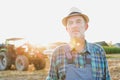 Senior farmer standing with arms corssed against farm tractor, round bale hay in field with yellow lens flare in background