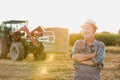 Senior farmer standing with arms corssed against farm tractor, round bale hay in field with yellow lens flare in background