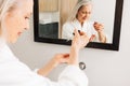 Senior female in a bathrobe drips liquid facial treatment on a palm in front of a mirror. Hands of an aged woman with a hyaluronic