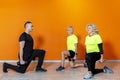 Senior fit couple doing weight lifting workout exercise standing at the gym isolated on orange background with personal trainer. Royalty Free Stock Photo