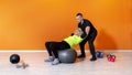 Senior fit woman doing workout exercise siting on a ball at the gym isolated on orange background followed by a pesonal trainer. Royalty Free Stock Photo