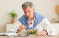 Senior Gentleman Eating Salad Reading News On Tablet Sitting Indoor