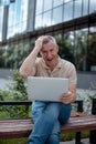 Senior glad gray haired businessman sitting on bench near building on city street and holding digital laptop. Modern