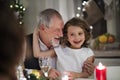 Senior grandfather with small granddaughter indoors at Christmas, sitting at table.