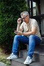 Senior gray haired businessman sitting in glasses on stairs near house and browsing on smartphone. Modern lifestyle Royalty Free Stock Photo