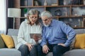 Senior gray-haired couple man and woman sitting on sofa at home. use a tablet computer for video communication and online Royalty Free Stock Photo