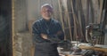 Senior gray haired male carpenter standing at wood manufacture with crossed arms smiling positively into camera.