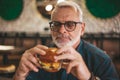 Senior gray-haired man in glasses with a beard is eating an appetizing burger. Lunch break, rest in the pub