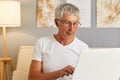 Senior gray haired man wearing white T-shier and glasses sitting on couch in living room holding laptop typing on keyboard looking Royalty Free Stock Photo