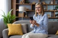 Senior gray-haired woman sitting joyfully on sofa at home, pensioner holding phone in hands, using app on smartphone Royalty Free Stock Photo