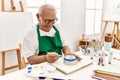 Senior grey-haired artist man smiling happy painting pottery sitting on the table at art studio Royalty Free Stock Photo