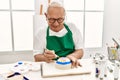 Senior grey-haired artist man smiling happy painting pottery sitting on the table at art studio Royalty Free Stock Photo