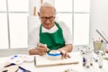 Senior grey-haired artist man smiling happy painting pottery sitting on the table at art studio Royalty Free Stock Photo