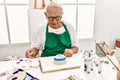 Senior grey-haired artist man smiling happy painting pottery sitting on the table at art studio Royalty Free Stock Photo