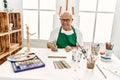 Senior grey-haired artist man smiling happy painting sitting on the table at art studio Royalty Free Stock Photo