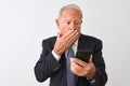 Senior grey-haired businessman using smartphone standing over isolated white background cover mouth with hand shocked with shame Royalty Free Stock Photo