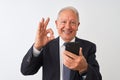 Senior grey-haired businessman using smartphone standing over isolated white background doing ok sign with fingers, excellent Royalty Free Stock Photo