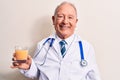 Senior grey-haired doctor man wearing stethoscope drinking glass of healthy orange juice looking positive and happy standing and Royalty Free Stock Photo
