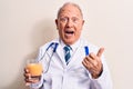 Senior grey-haired doctor man wearing stethoscope drinking glass of healthy orange juice pointing thumb up to the side smiling Royalty Free Stock Photo