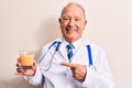 Senior grey-haired doctor man wearing stethoscope drinking glass of healthy orange juice smiling happy pointing with hand and Royalty Free Stock Photo