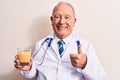 Senior grey-haired doctor man wearing stethoscope drinking glass of healthy orange juice smiling happy and positive, thumb up Royalty Free Stock Photo